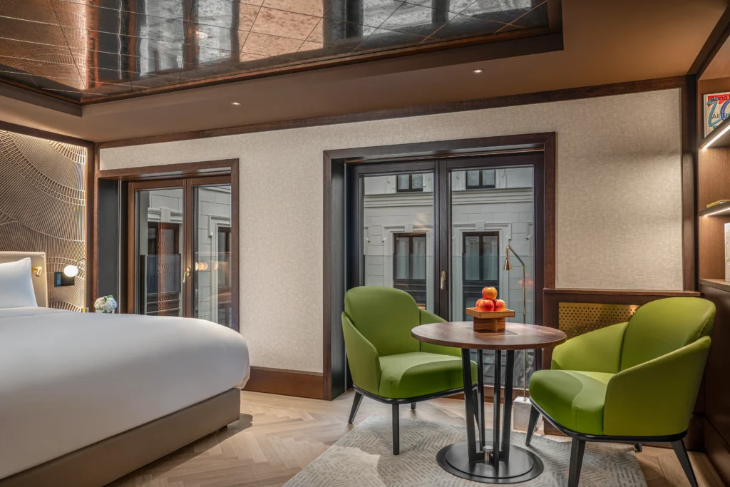 Modern hotel room with a large bed, two green armchairs around a small round table with fruit, and floor-to-ceiling windows facing another building. Our rooms and suites offer comfort and contemporary style for your stay.